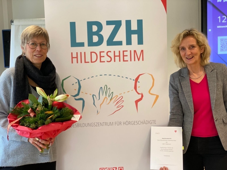 Vor einem Banner mit dem neuem Logo des LBZH Hildesheim stehen von links die Direktion, Frau Wüst und die Projektleiterin, Frau Berger-Nowak. Frau Wüst hält einen Blumenstrauß in der Hand, Frau Berger-Nowak präsentiert den Abschlussbericht