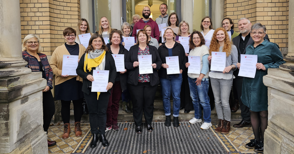 Zu sehen sind 19 Teilnehmende der Qualifizierungsmaßnahme „Deutsche Gebärdensprache“ sowie Frau Stasche als Vertretung des Trägerteams des Landesamtes für Soziales, Jugend und Familie und Herr Dr. Westerheide, Direktor des LBZH OS